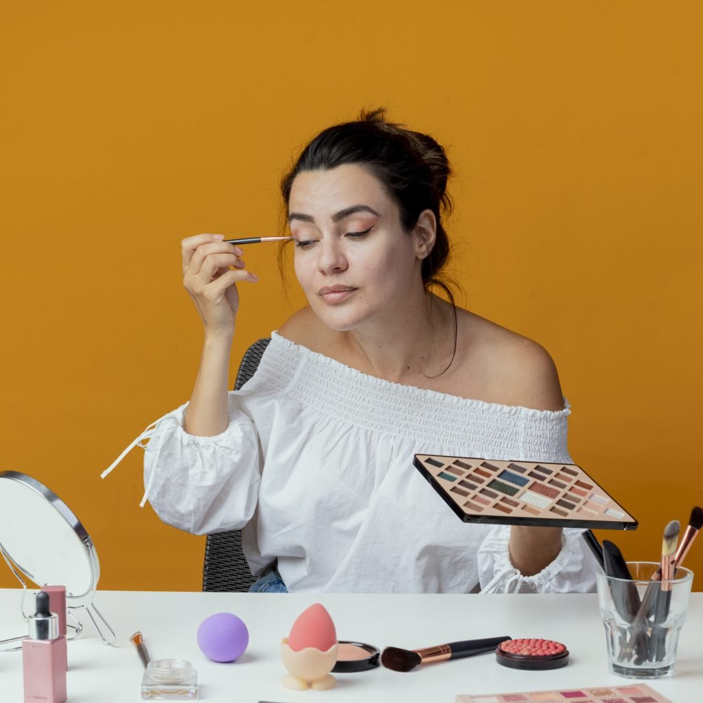 pleased beautiful girl sits at table with makeup tools looks at mirror applying eyeshadow with makeup brush isolated on orange background with copy space